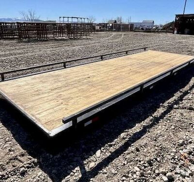 Empty wooden-deck flatbed trailer on a gravel lot, with metal cattle pens visible in the background.